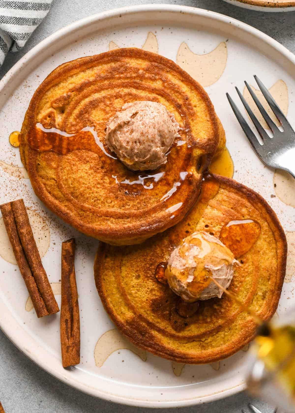 two Pumpkin pancakes on a white ceramic plate, each one is topped with a scoop of cinnamon honey butter, a drizzle of maple syrup and a sprinkle of cinnamon,. There are two cinnamon sticks and a fork on the plate as well.