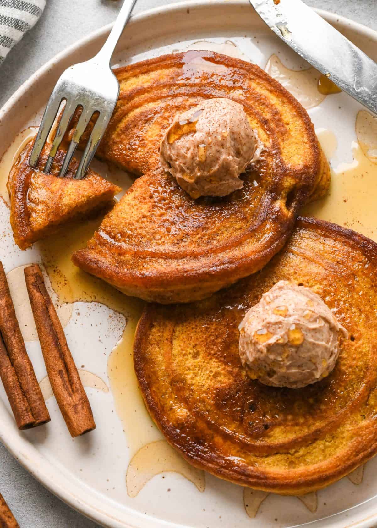 two Pumpkin pancakes on a white ceramic plate, each one is topped with a scoop of cinnamon honey butter and maple syrup. There are two cinnamon sticks and a fork with a bite on it on the plate as well.