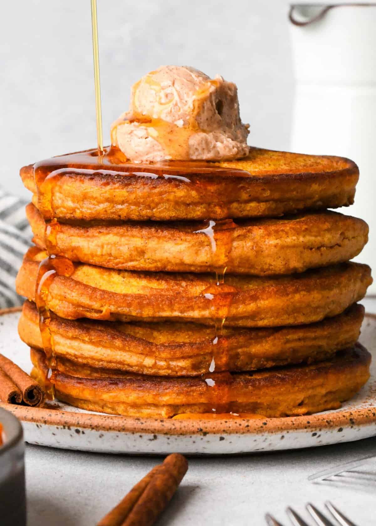 a stack of 5 pumpkin pancakes on a white ceramic plate topped with a scoop of cinnamon butter with maple syrup on top and dripping down the sides. There are two cinnamon sticks, a bowl of pumpkin puree and a striped towel around the plate