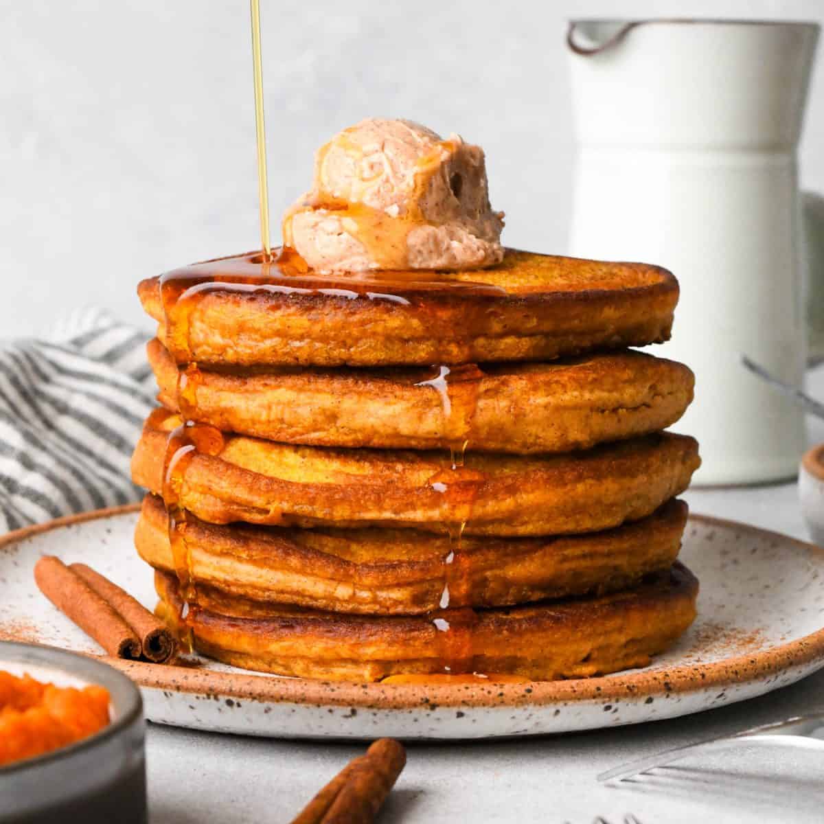 syrup being poured over a stack of 5 pumpkin pancakes on a white ceramic plate topped with a scoop of cinnamon butter with maple syrup on top and dripping down the sides. There are two cinnamon sticks, a bowl of pumpkin puree and a striped towel around the plate
