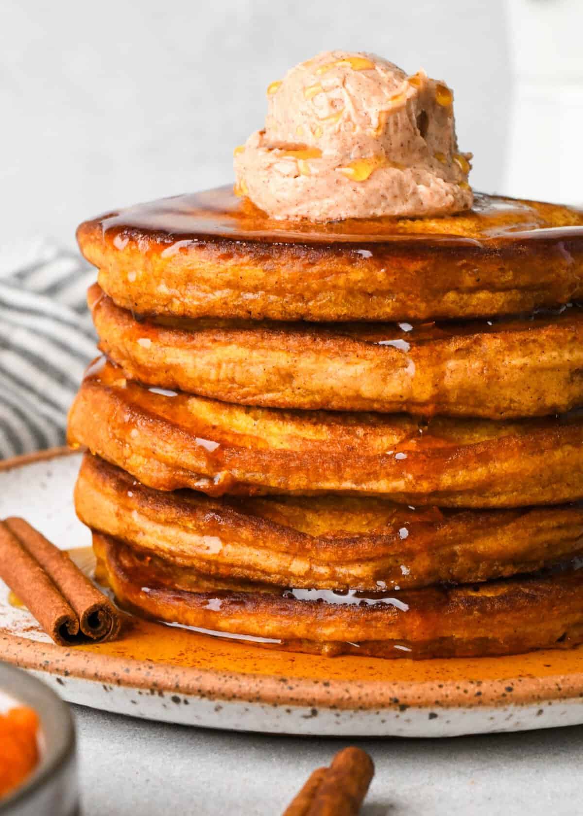 a stack of 5 pumpkin pancakes on a white ceramic plate topped with a scoop of cinnamon butter with maple syrup on top and dripping down the sides. There are two cinnamon sticks, a bowl of pumpkin puree and a striped towel around the plate