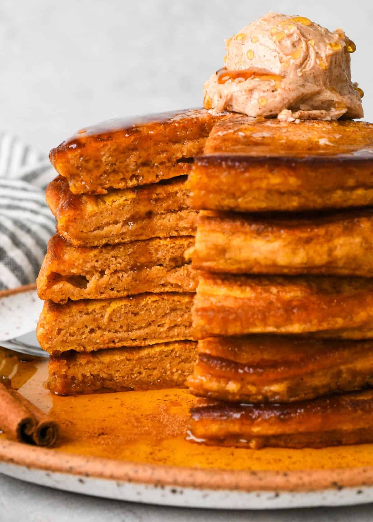 close up photo of a stack of 5 moist and fluffy pumpkin pancakes topped with a scoop of cinnamon honey butter and drizzled with maple syrup, a bite is cut out of the stack (all 5 pancakes) so the moist and fluffy inside is visible.