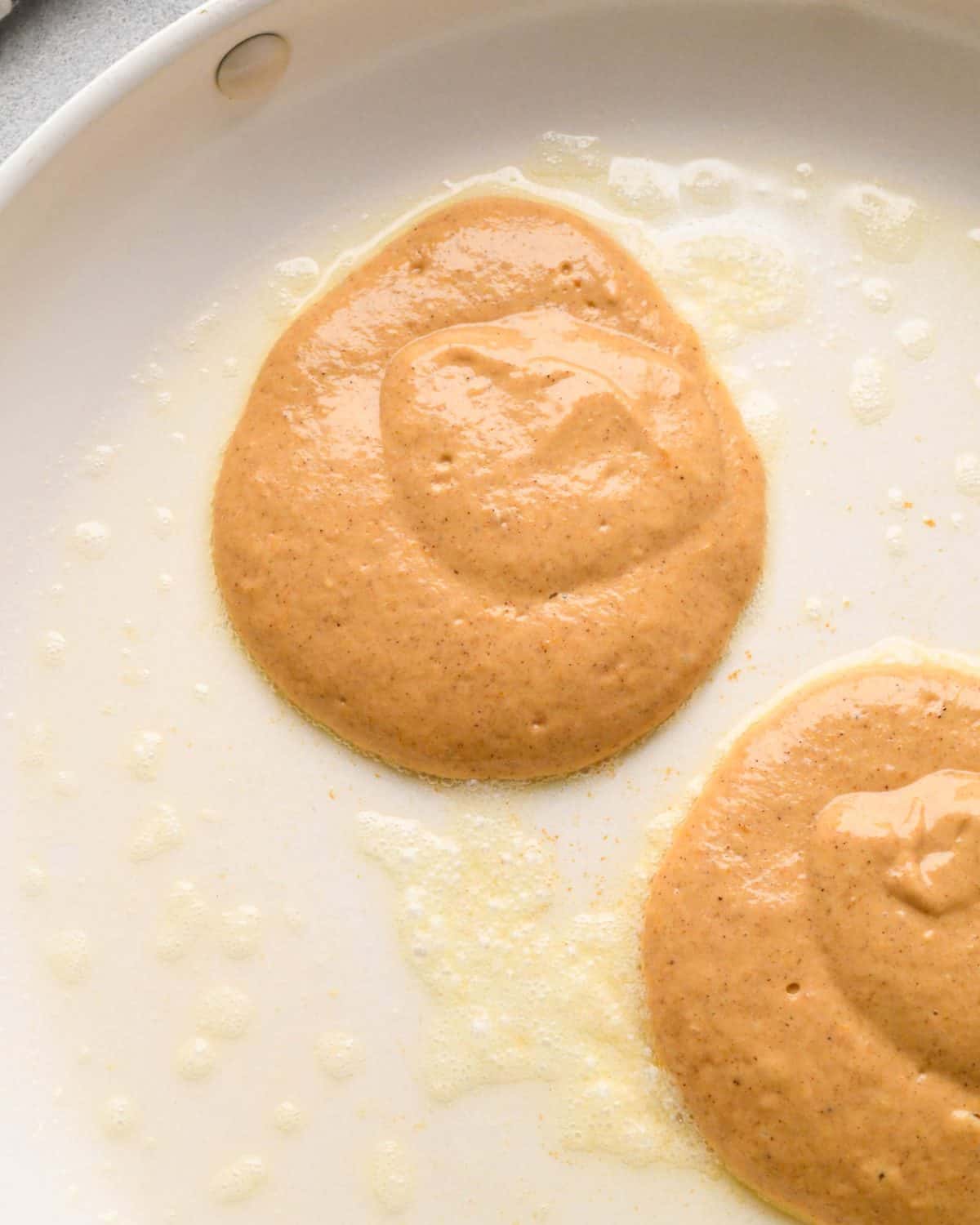 2 pumpkin pancakes cooking in melted butter on the first side on a white nonstick frying pan