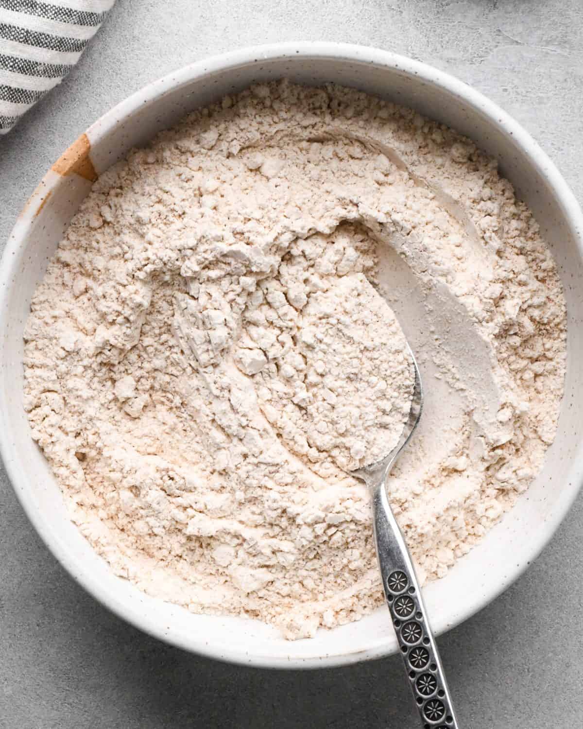 making pumpkin pancakes recipe - flour, baking powder, baking soda, salt, cinnamon and pumpkin pie spice in a small white ceramic bowl after mixing with a silver spoon