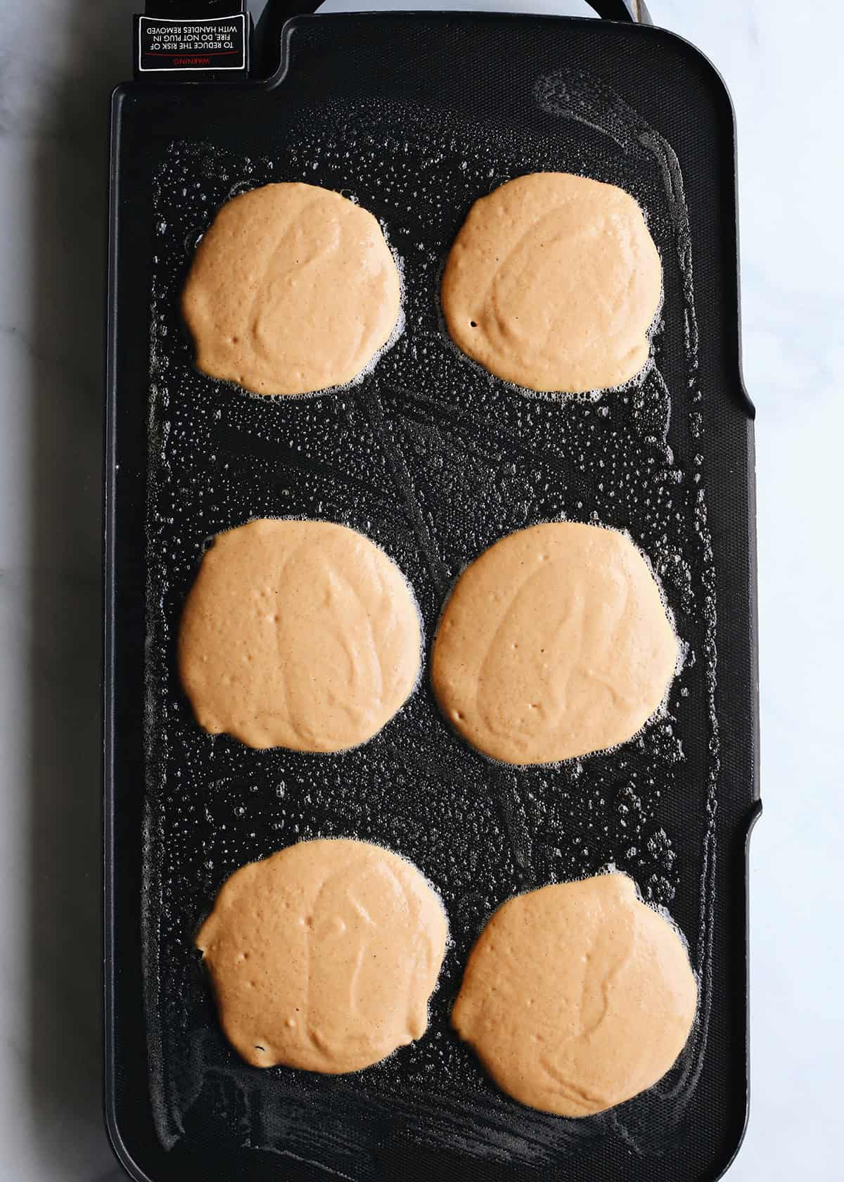 6 pumpkin pancakes cooking in melted butter on the first side on a black electric griddle