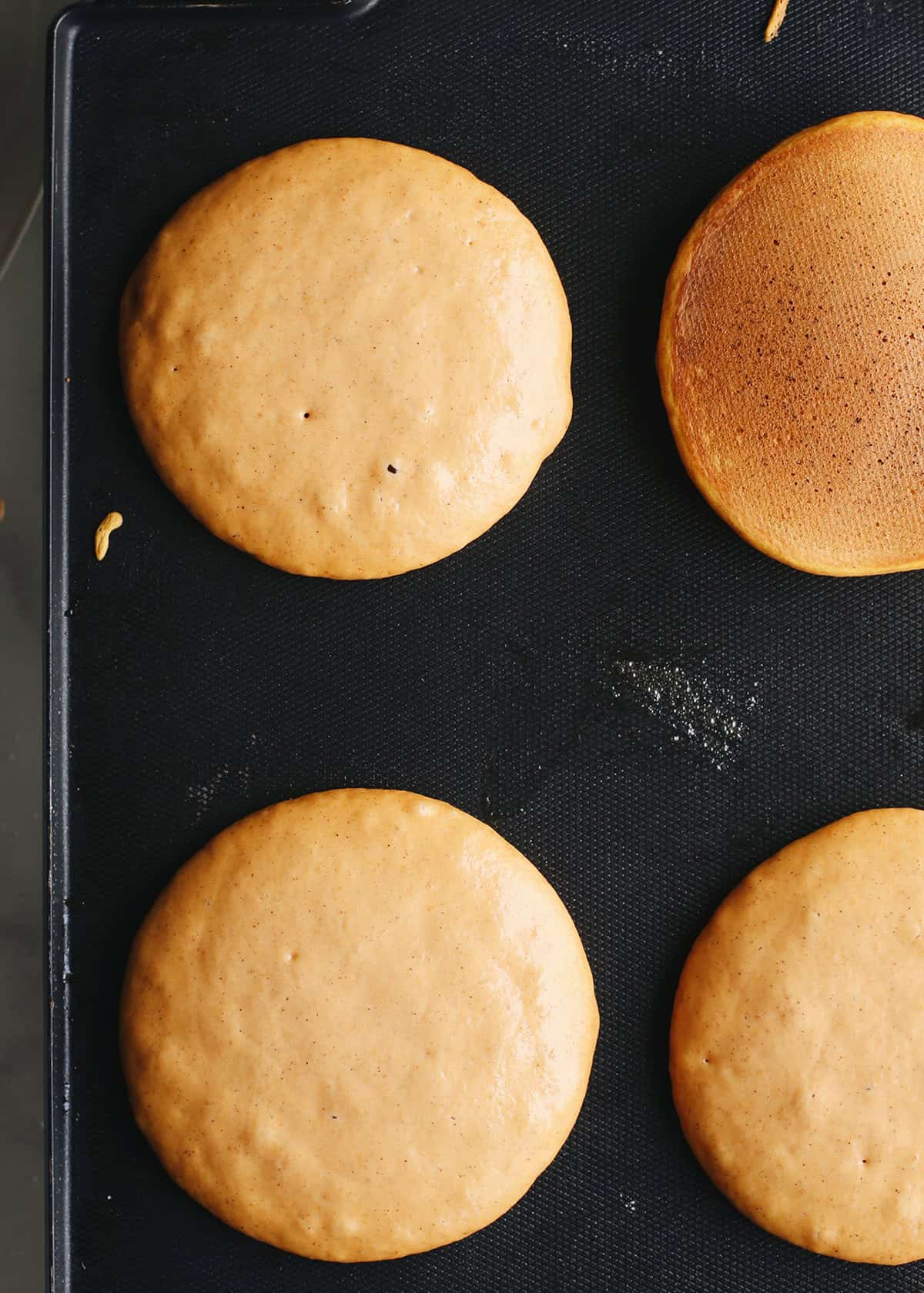 4 pumpkin pancakes cooking in melted butter on a black electric griddle, bubbles are visible in the batter indicating it's time to flip the pancakes, one in the top right corner has already been flipped.