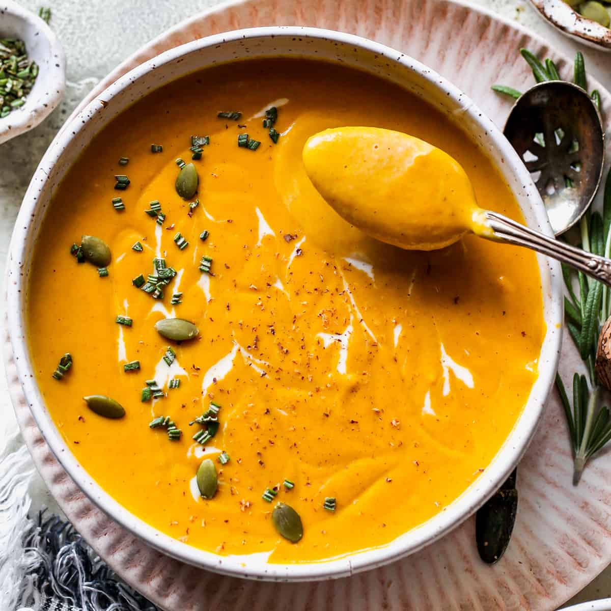 a spoon taking a scoop out of a bowl of Sweet Potato Soup garnished with rosemary, cream and pepitas. 
