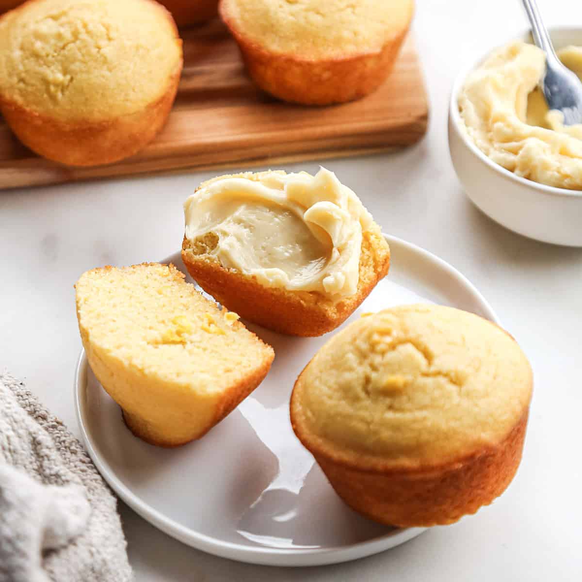 two Cornbread Muffins on a plate, one cut in half with butter spread on one half