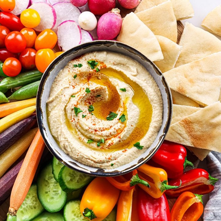 overhead view of a bowl of Homemade Hummus topped with olive oil, paprika and parsley surrounded by vegetables and pita bread