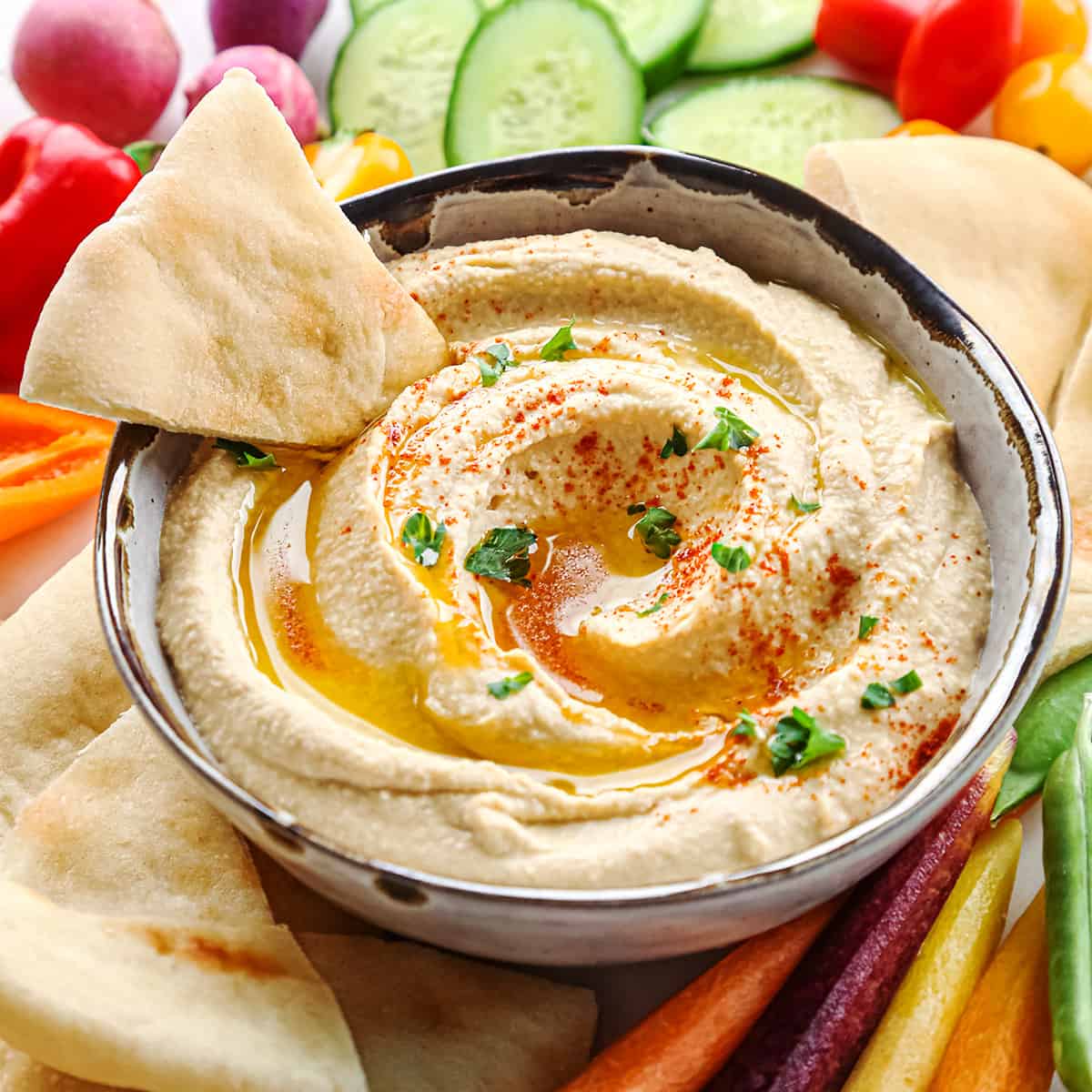a triangle of pita bread being dipped into a bowl of Homemade Hummus topped with olive oil, paprika and parsley