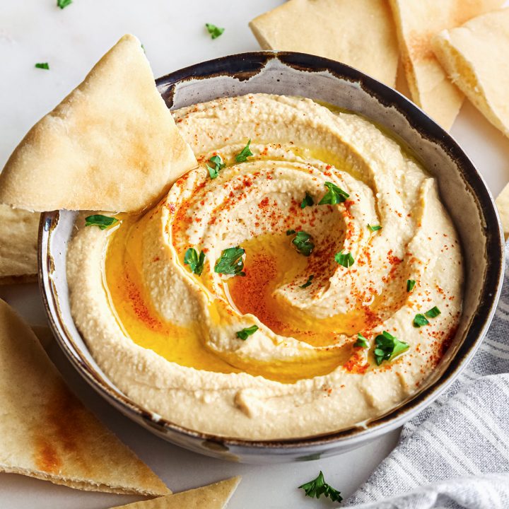 a triangle of pita bread being dipped into a bowl of Homemade Hummus topped with olive oil, paprika and parsley