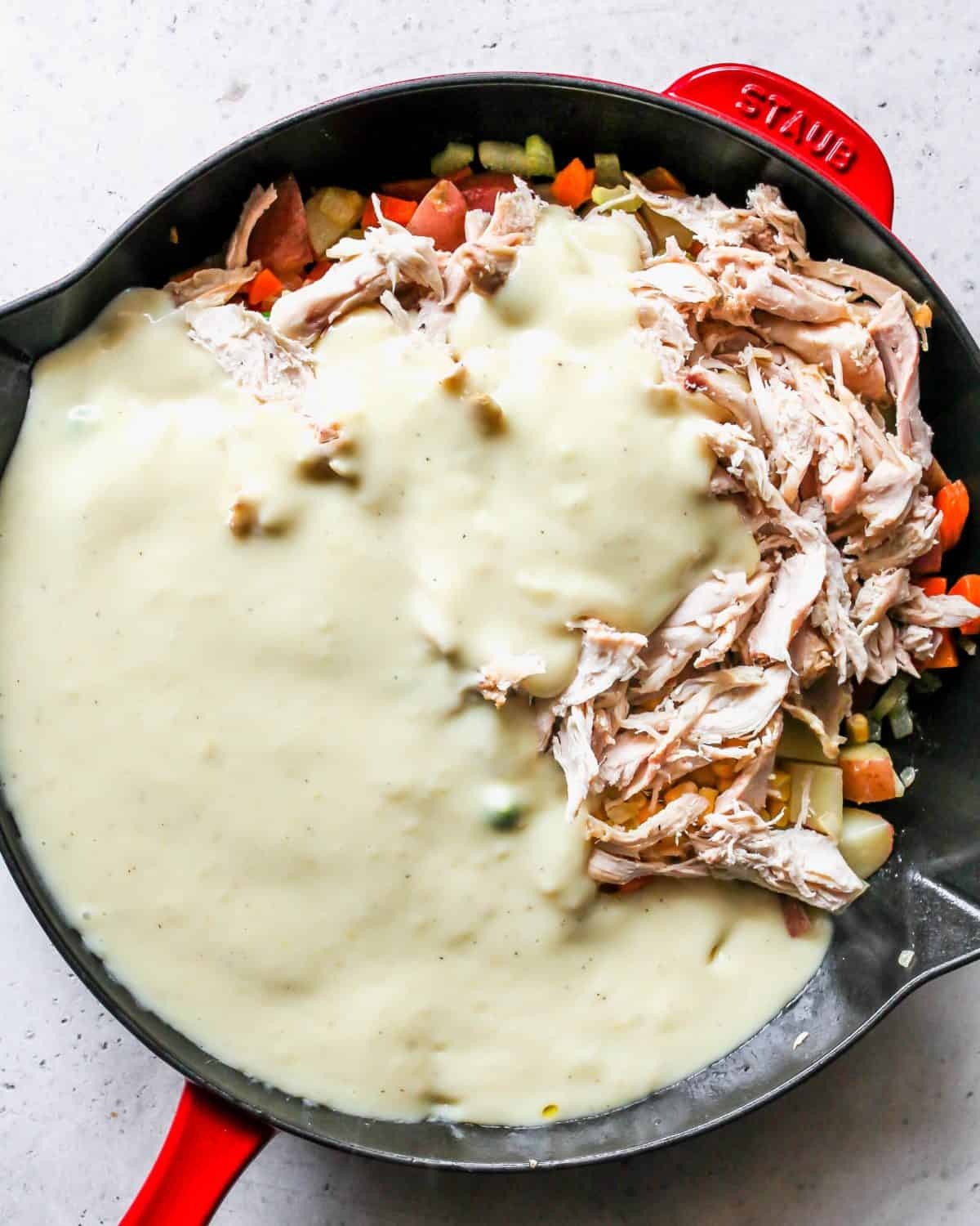 making turkey pot pie recipe - sauce added to the filling ingredients in a large cast iron skillet with a black bottom and red exterior.