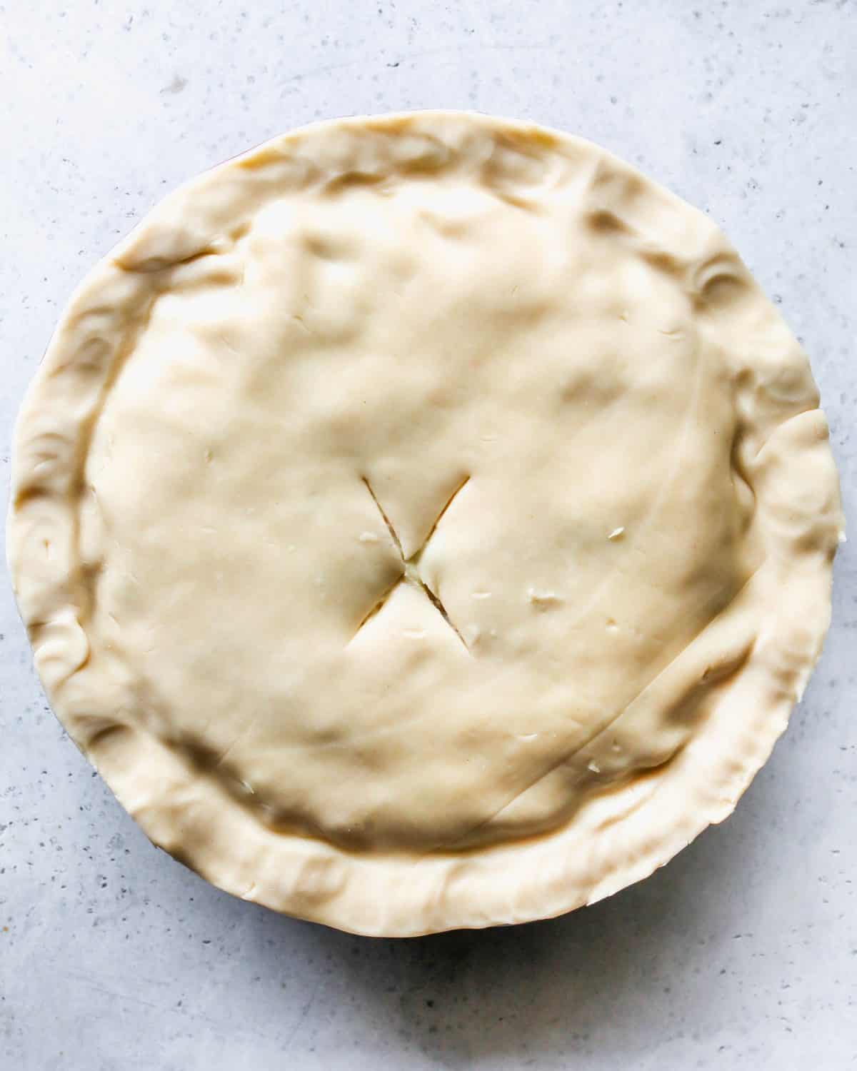 assembled turkey pot pie with pinched edges and an "X" cut into the top pie crust before baking on a gray surface