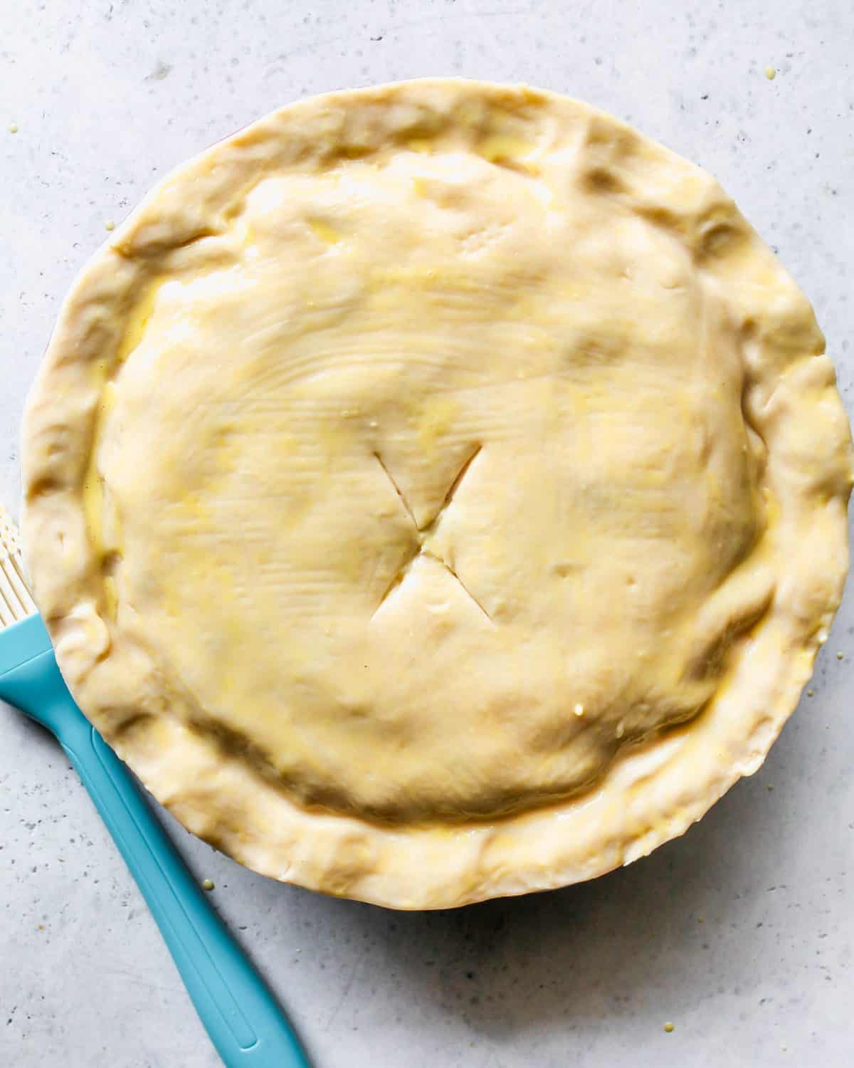 assembled turkey pot pie with pinched edges and an "X" cut into the top pie crust before baking on a gray surface with egg wash brushed over it.