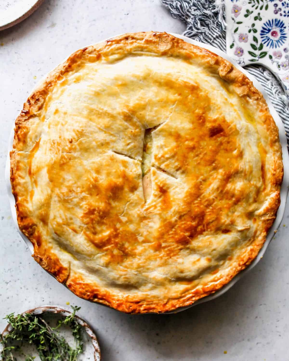 baked turkey pot pie in a white baking dish with a bowl of thyme in the bottom left corner and a towel and pie server in the top right corner