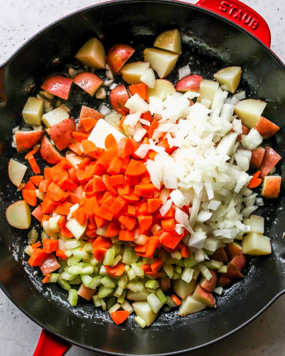 making turkey pot pie recipe - red potatoes, minced garlic, onion, red pepper, celery and butter cooking in a large cast iron skillet with a black bottom and red exterior.