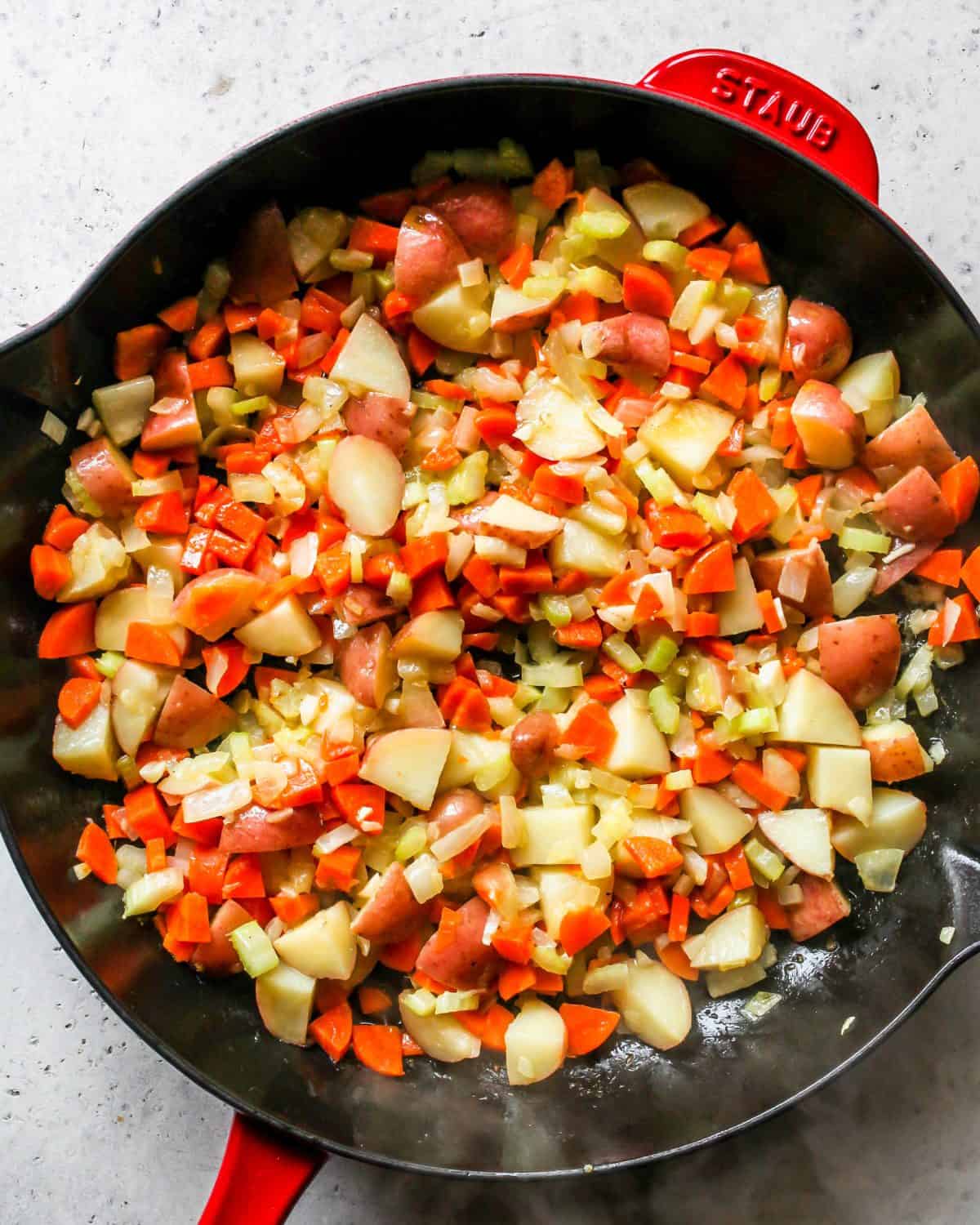 making turkey pot pie recipe - red potatoes, minced garlic, onion, red pepper, celery and butter cooking in a large cast iron skillet with a black bottom and red exterior.