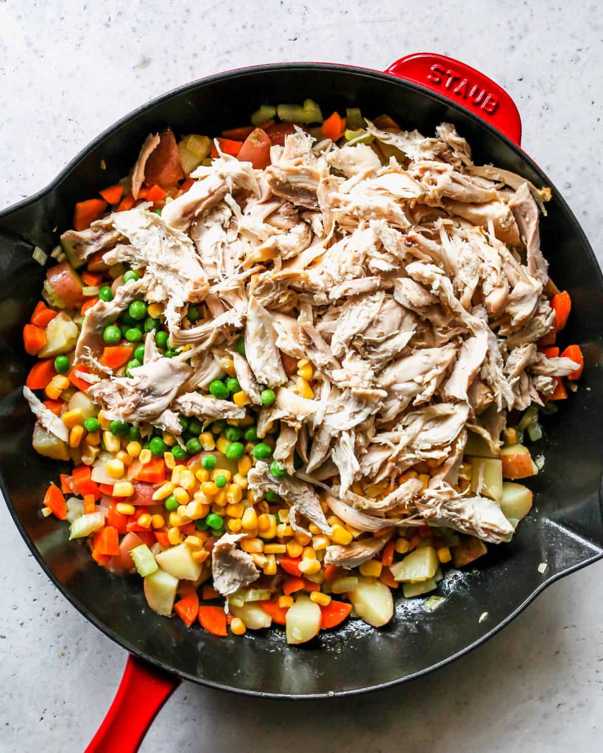 making turkey pot pie recipe - turkey peas and corn added to the filling ingredients in a large cast iron skillet with a black bottom and red exterior.