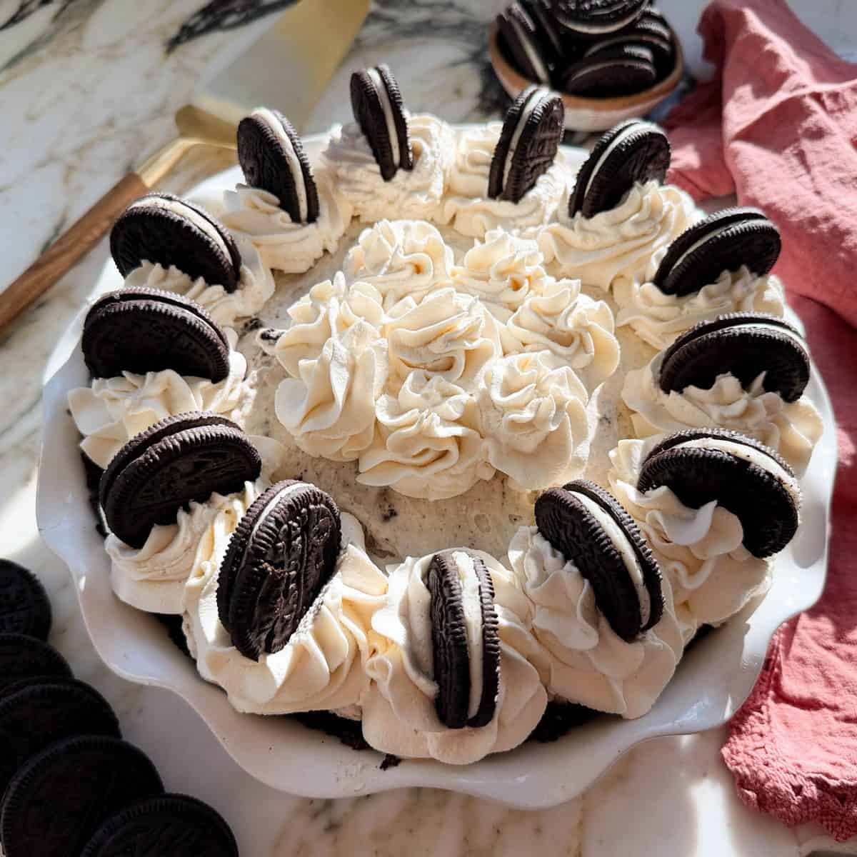 oreo pie recipe in a white cearmic pie dish topped with whipped cream and oreos. There are oreos and a pie server in the background.