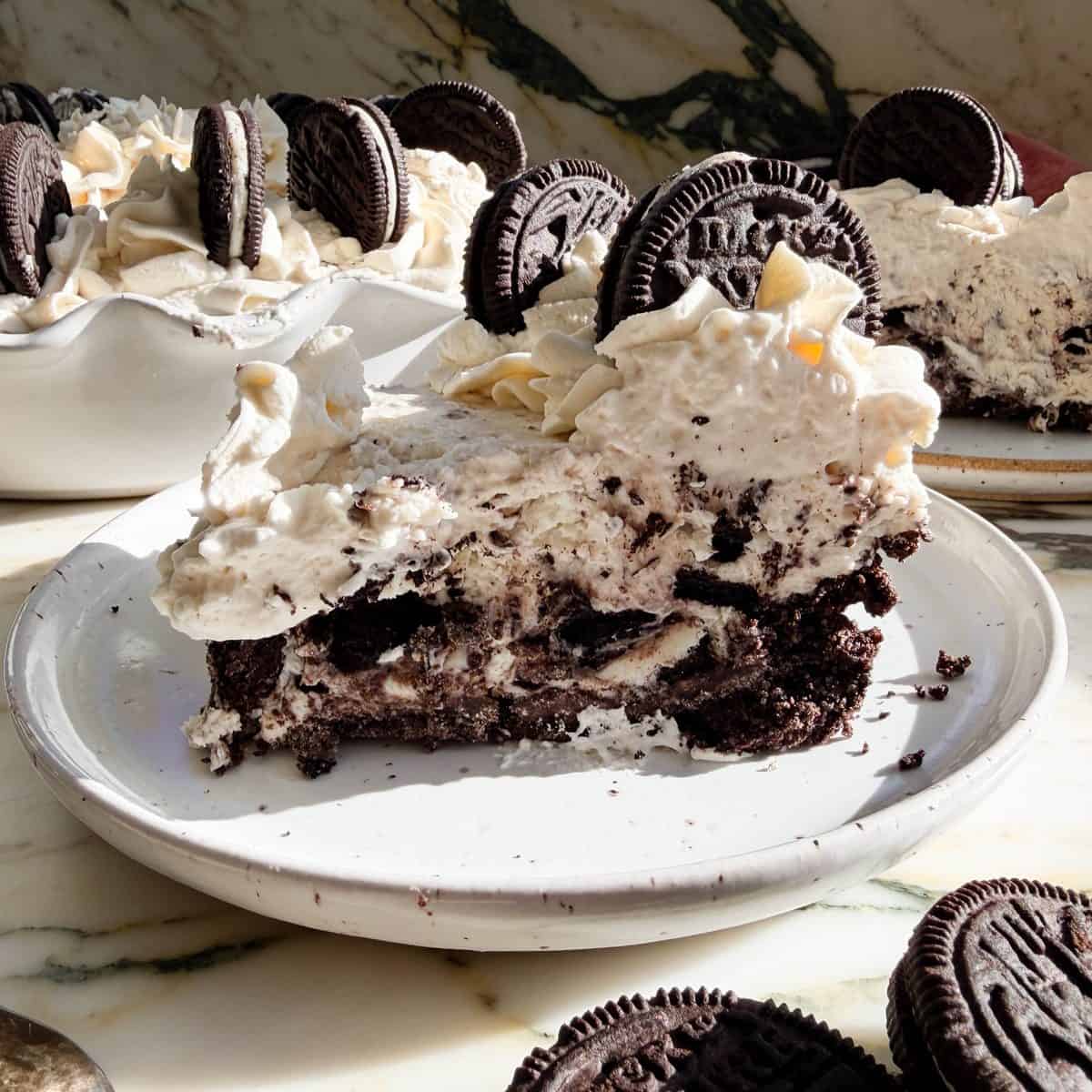 a slice of oreo pie on a with ceramic plate topped with whipped cream and two Oreo cookies. there is another slice of pie and the whole pie in the background.