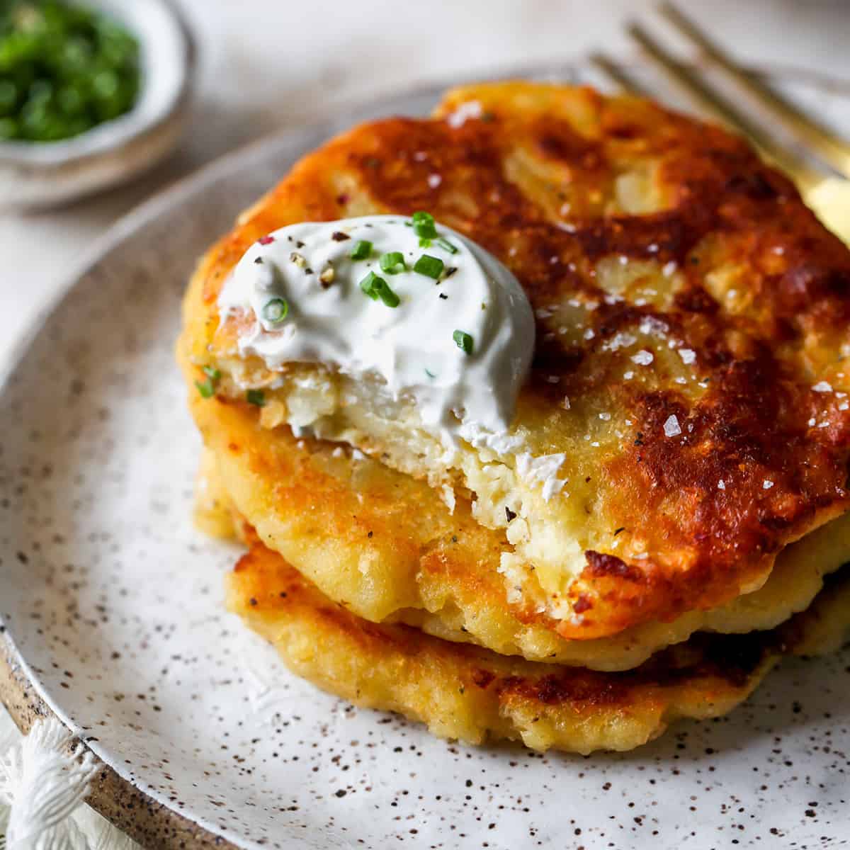 a stack of 3 Potato Pancakes, the top one has a bite taken out of it. Garnished with sour cream and chives.