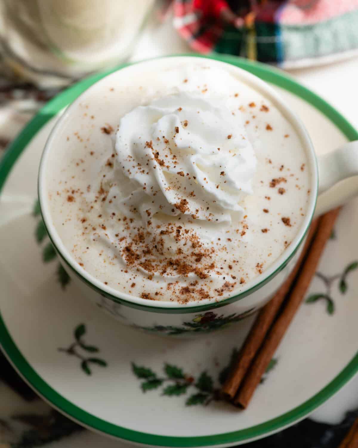 overhead photo of homemade eggnog recipe in a Christmas tree mug topped with whipped cream and a sprinkle of nutmeg on a saucer with a cinnamon stick on the saucer and a christmas towel in the background