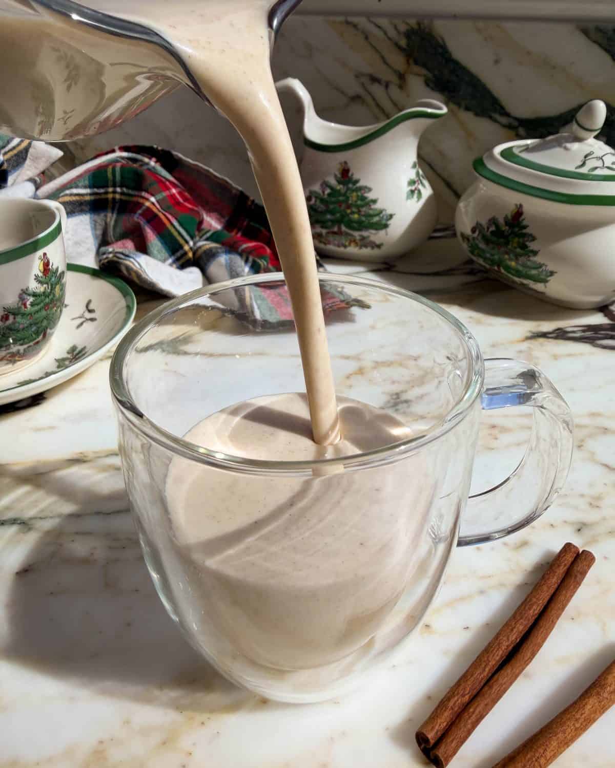homemade eggnog being poured into a glass mug so it's thick and creamy texture is visible. There is a Christmas mug and other Christmas dishes int he background and two cinnamon sticks in the bottom right corner.