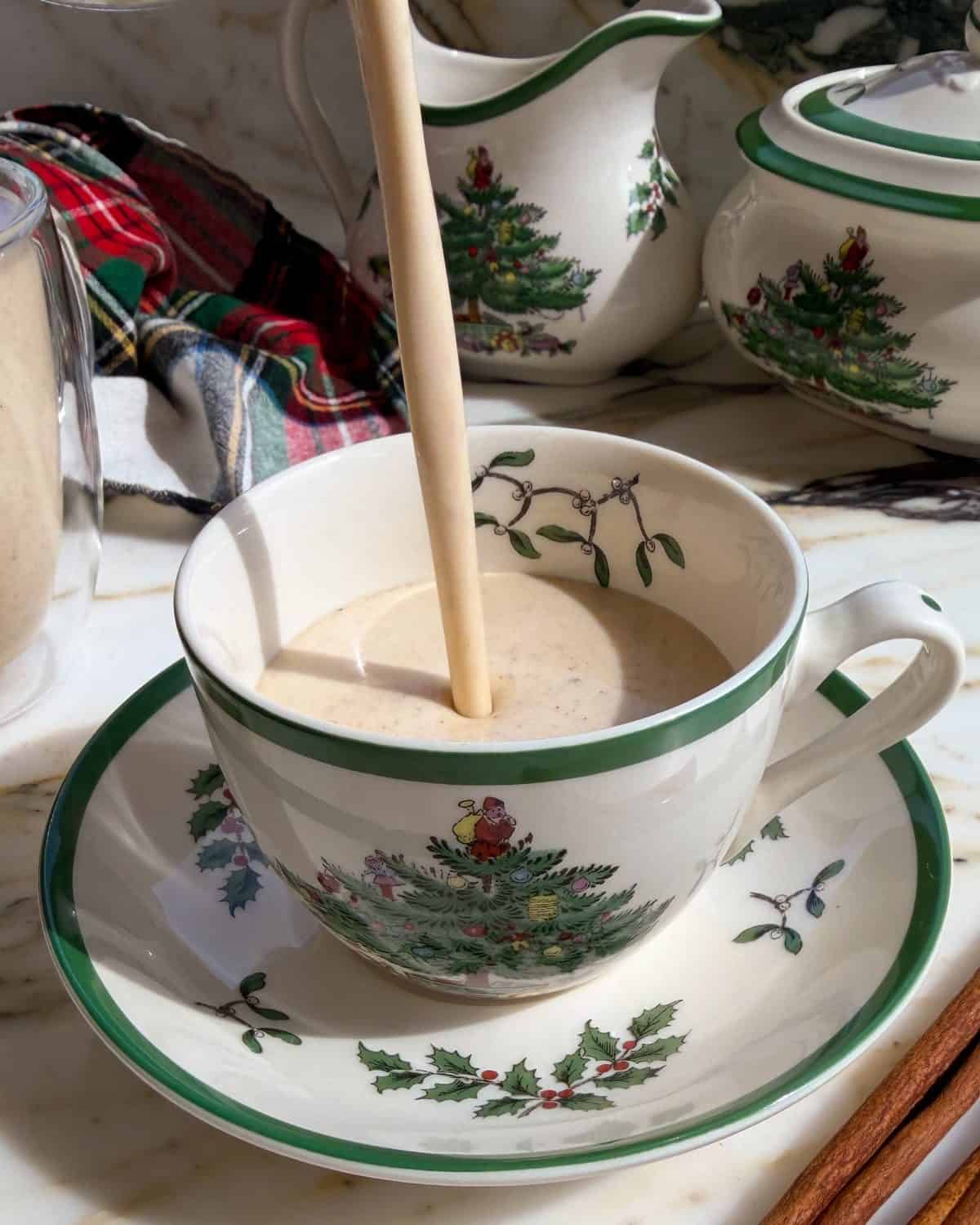 homemade eggnog recipe being poured into a small Christmas tree tea cup on a saucer so the thick creamy texture is visible with another glass mug of eggnog in the background and other Christmas tree dishes.