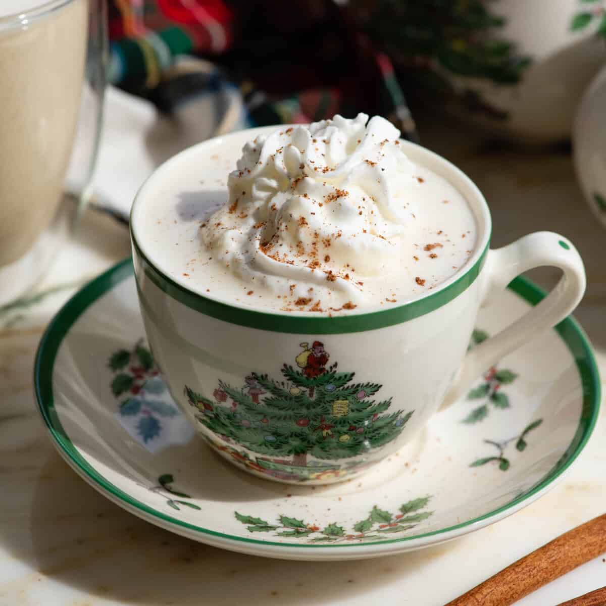 homemade eggnog recipe in a Christmas tree mug topped with whipped cream and a sprinkle of nutmeg on a saucer with another glass mug of eggnog and a Christmas towel in the background and a cinnamon stick in the bottom right corner