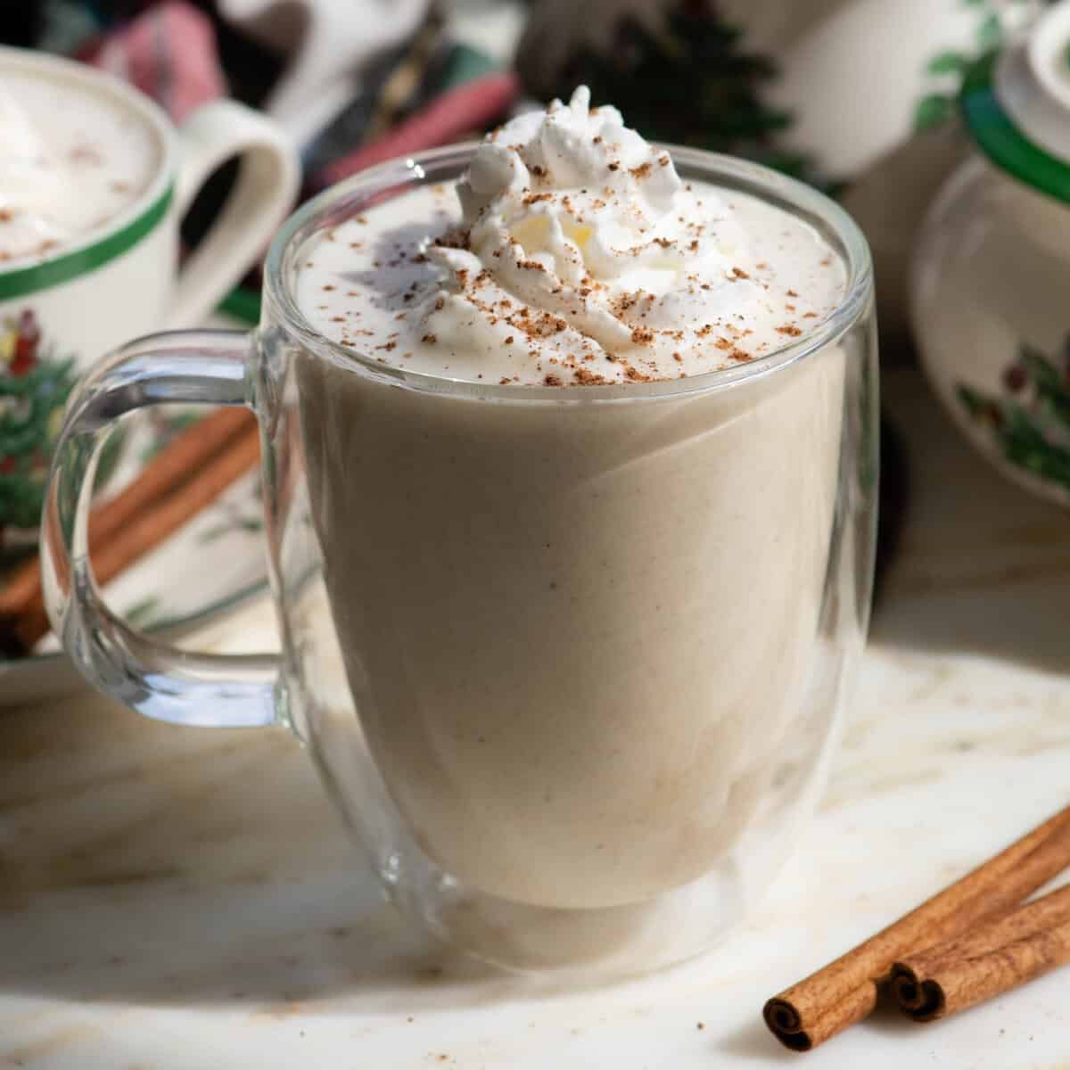 homemade eggnog recipe in a glass mug topped with whipped cream and a sprinkle of nutmeg with another mug of eggnog in the background and Christmas dishes and a cinnamon stick
