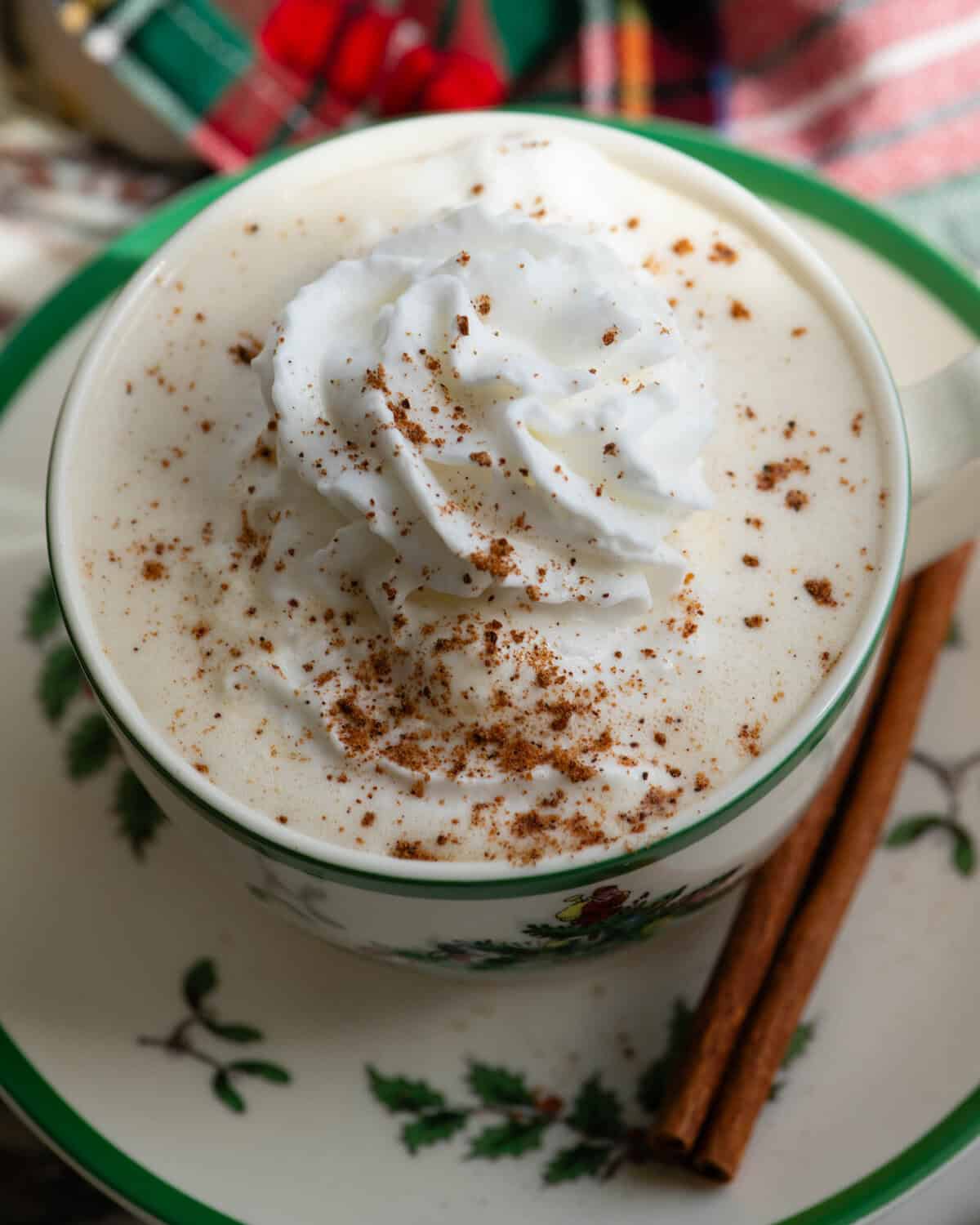 close up overhead photo of homemade eggnog recipe in a Christmas tree mug topped with whipped cream and a sprinkle of nutmeg on a saucer with a cinnamon stick on the saucer and a christmas towel in the background