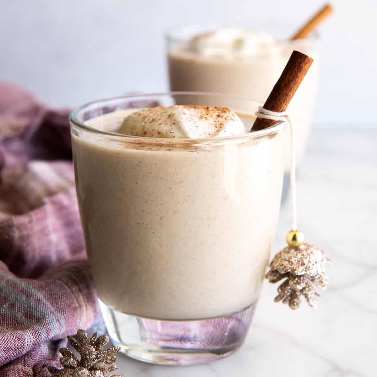 two glasses of Homemade Eggnog with whipped cream and ground nutmeg on top and a cinnamon stick inside.