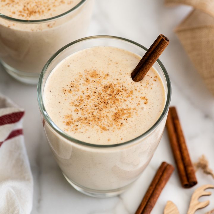 two glasses of Eggnog with a cinnamon stick in it and ground nutmeg on top