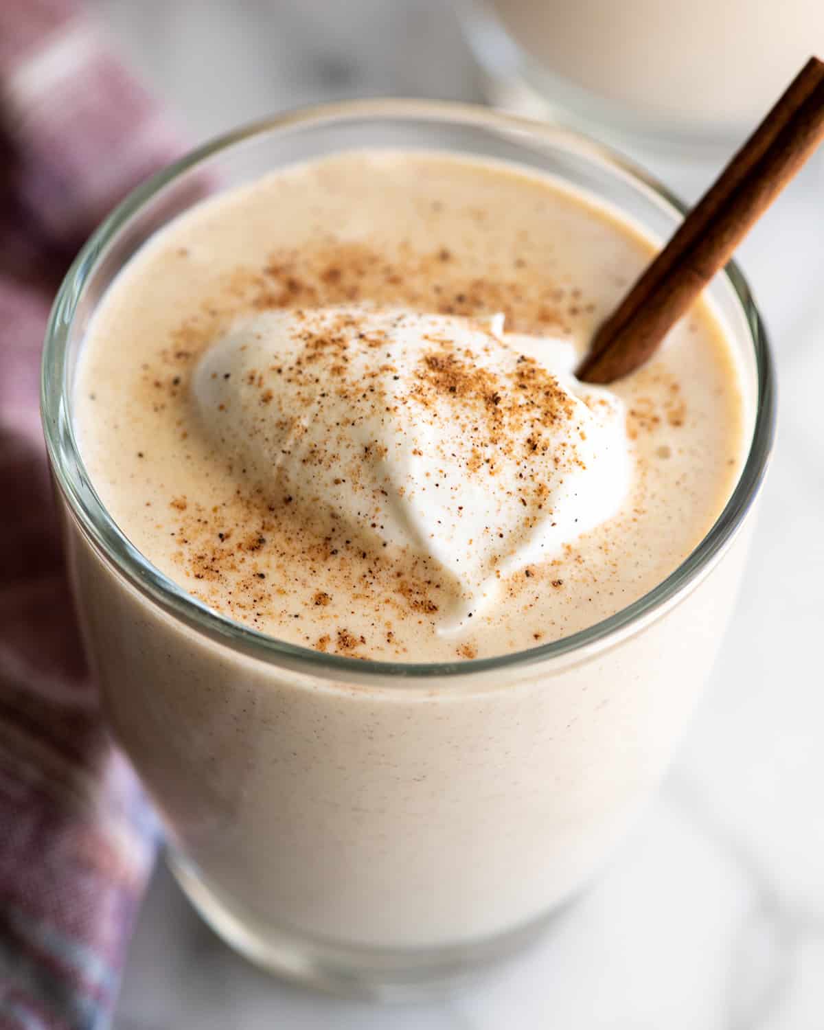a glass of Homemade Eggnog with whipped cream and ground nutmeg on top and a cinnamon stick inside.