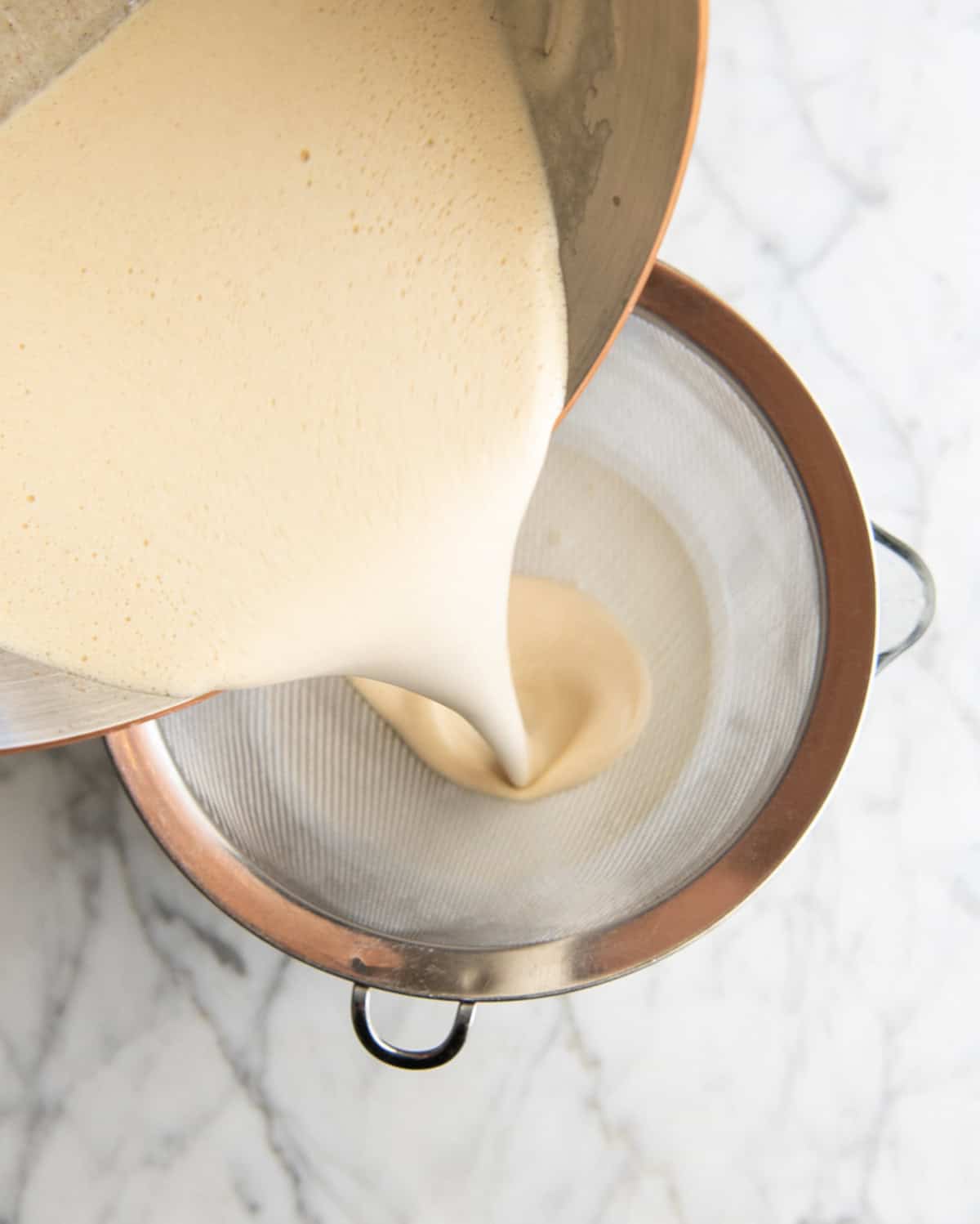 how to make eggnog recipe - custard mixture being poured into the metal strainer on the bowl with the heavy cream to be strained as it's added to the cream