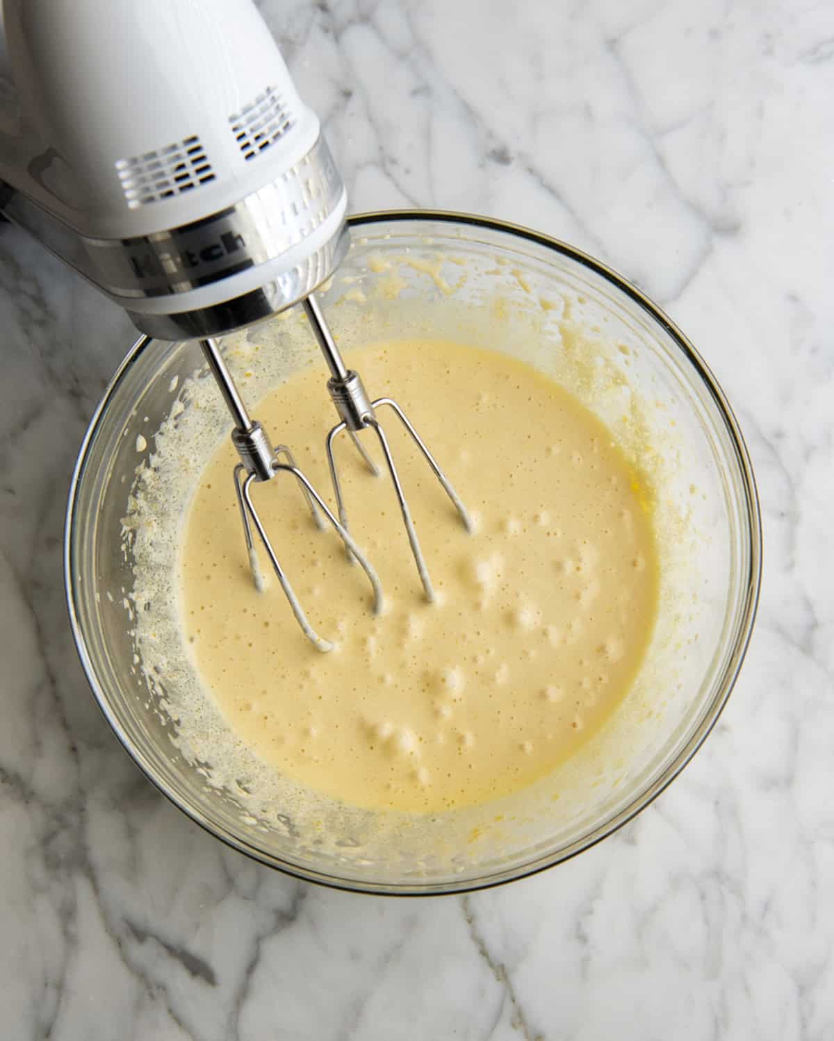 how to make eggnog recipe - yolk and sugar mixture in a glass mixing bowl with a white handheld mixer in the bowl on a marble surface after the eggs and sugar have been beaten and are pale yellow and thick