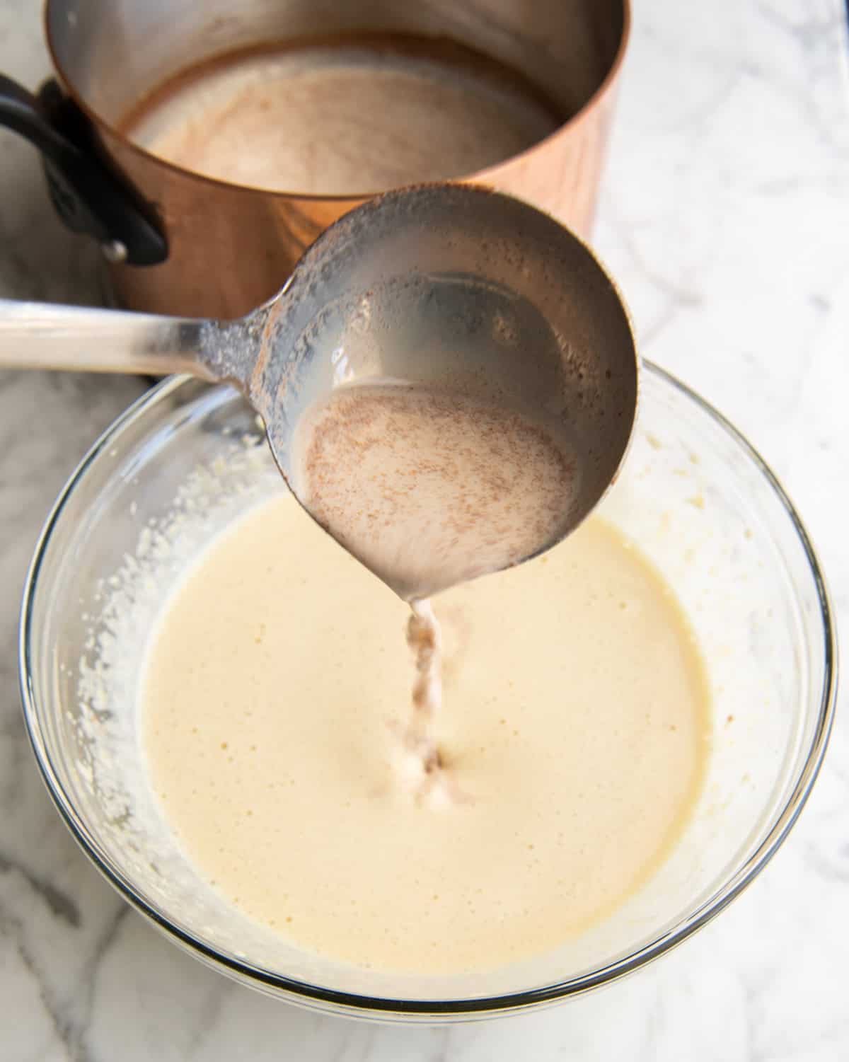 how to make eggnog recipe - warm milk/spice mixture being poured into the beaten eggs and sugar in a glass bowl with a metal ladle while the copper pot with the rest of the milk is in the background