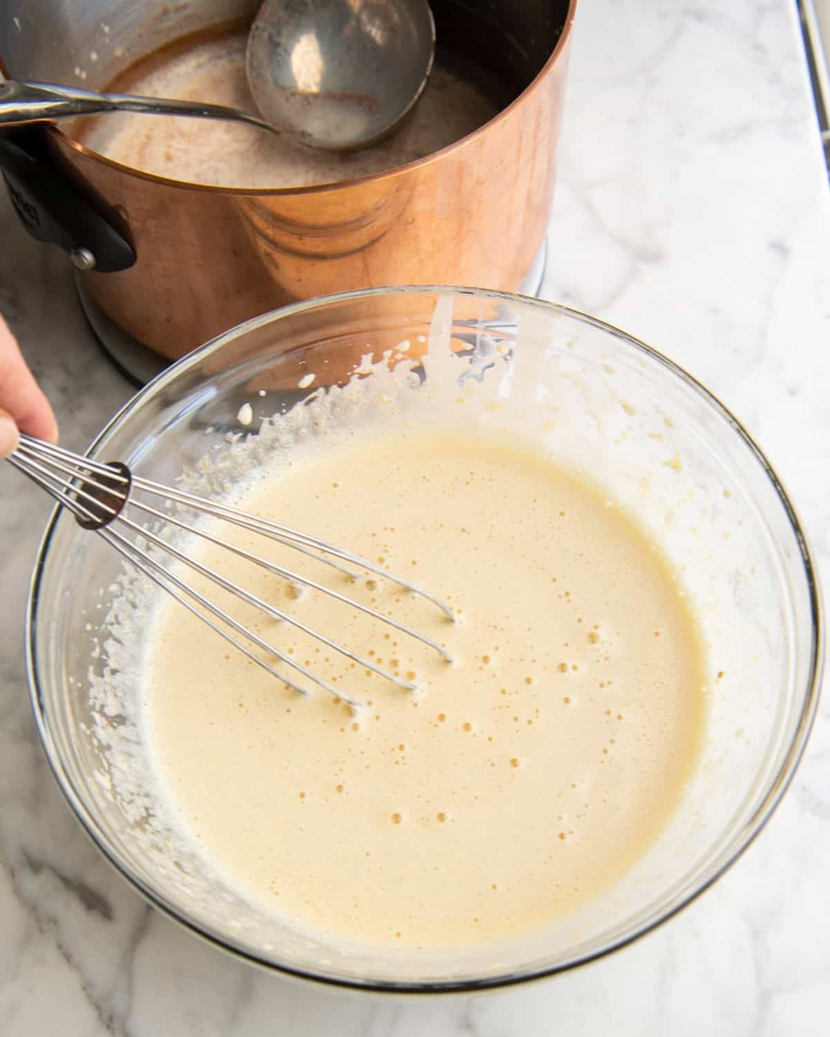 how to make eggnog recipe - warm milk/spice mixture being whisked into the beaten eggs and sugar in a glass bowl while the copper pot with the rest of the milk and a metal ladle is in the background