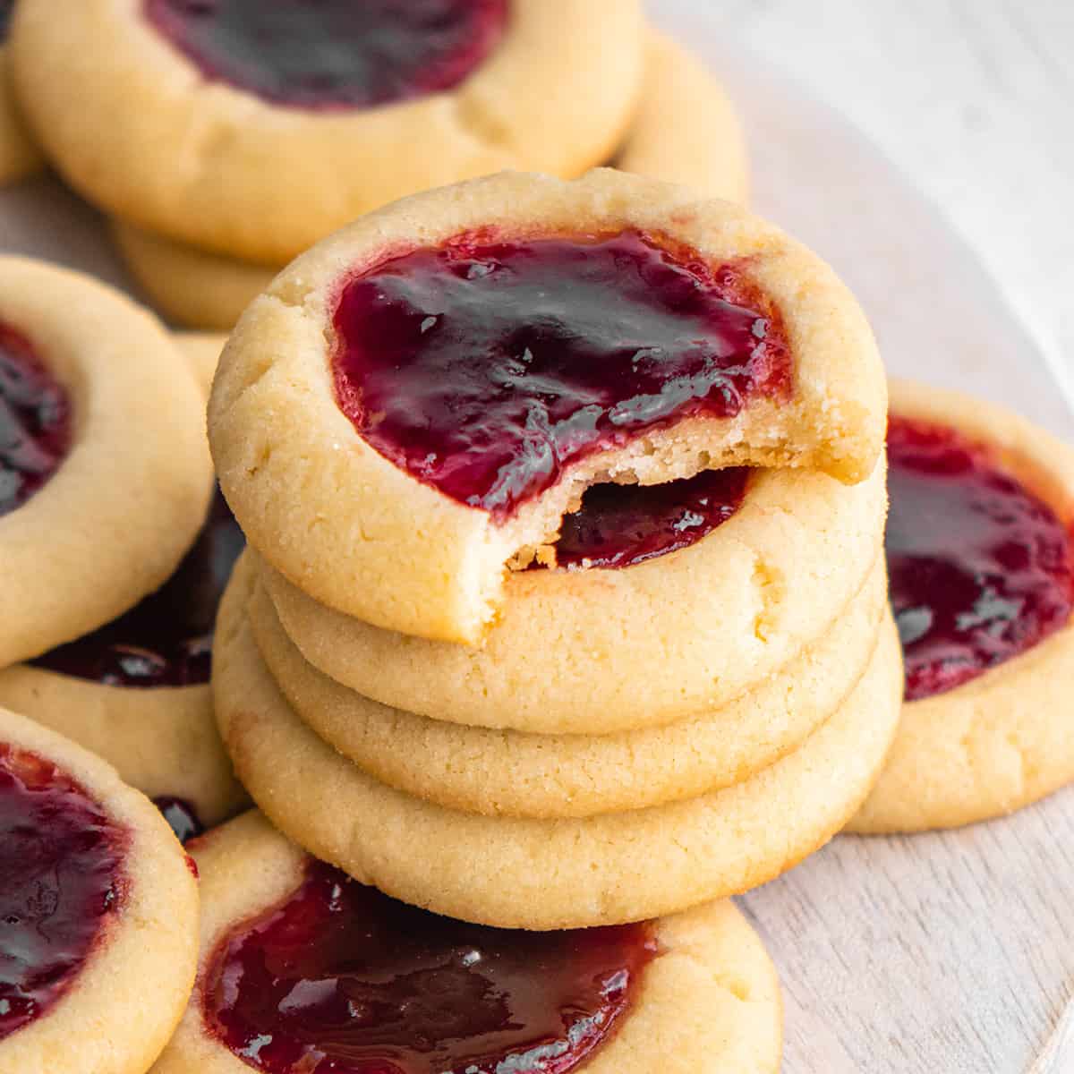 stack of four Thumbprint Cookies on top of other raspberry thumbprint cookies - top one has a bite taken out of it
