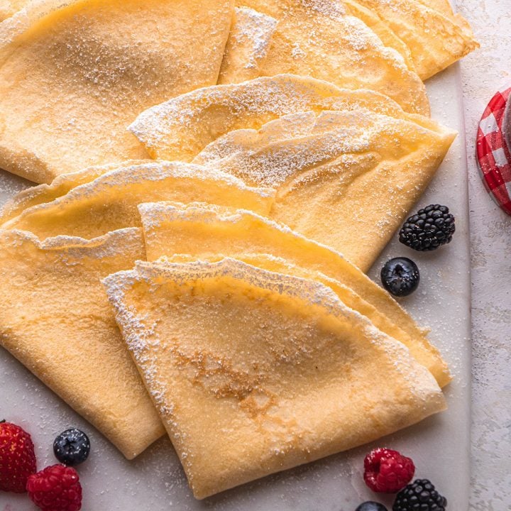6 crepes dusted with powdered sugar on a serving board with berries