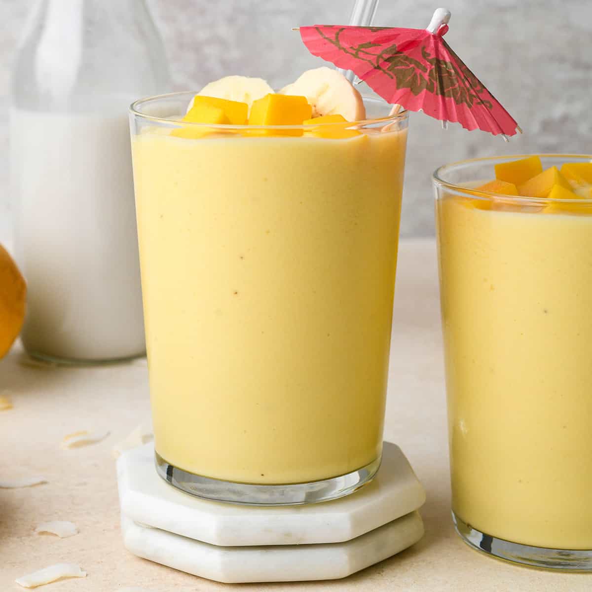 two glasses of Tropical Smoothie topped with banana and mango, one glass has a drink umbrella in it
