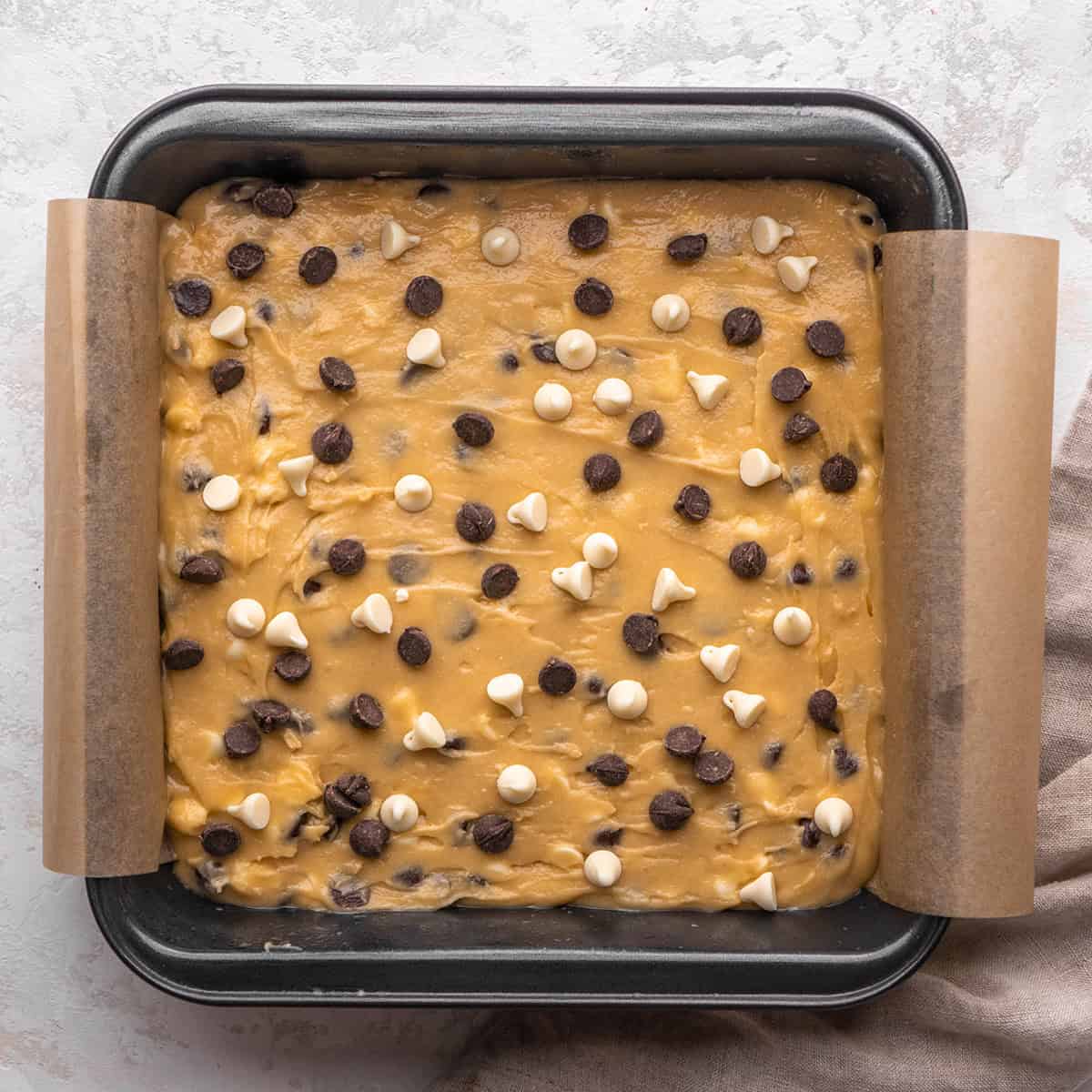 how to make blondies - batter spread into a square metal baking dish lined with parchment paper with white and semisweet chips on top before baking