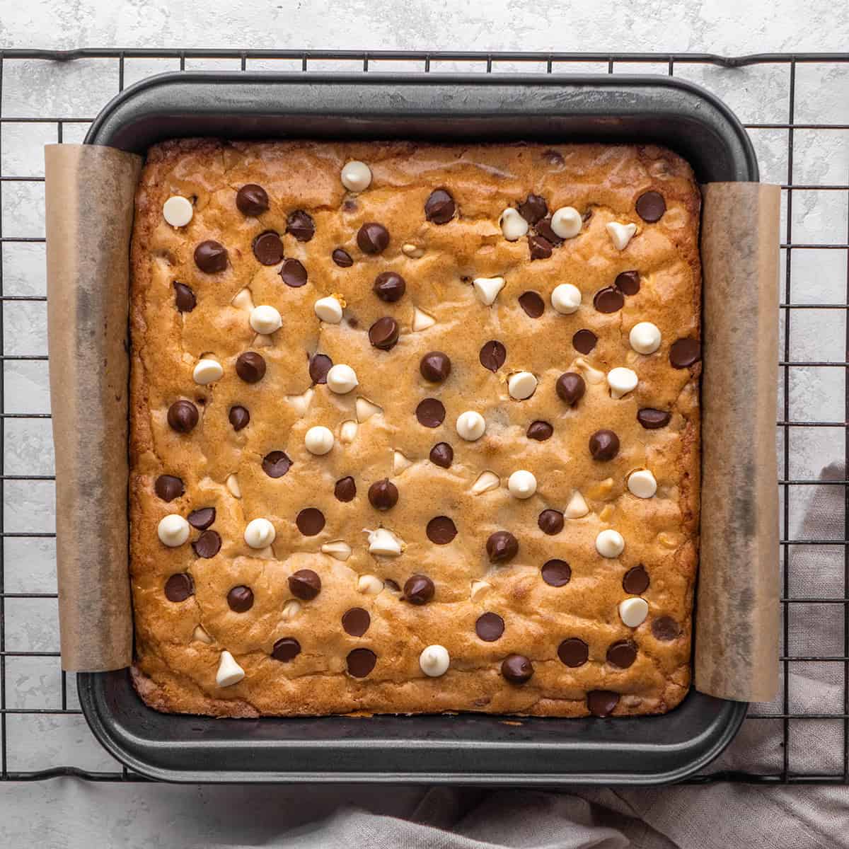 blondies recipe in a square metal baking dish lined with parchment paper on a wire cooling rack after baking. They have a golden brown, shiny top studded with lots of white and semisweet chips.