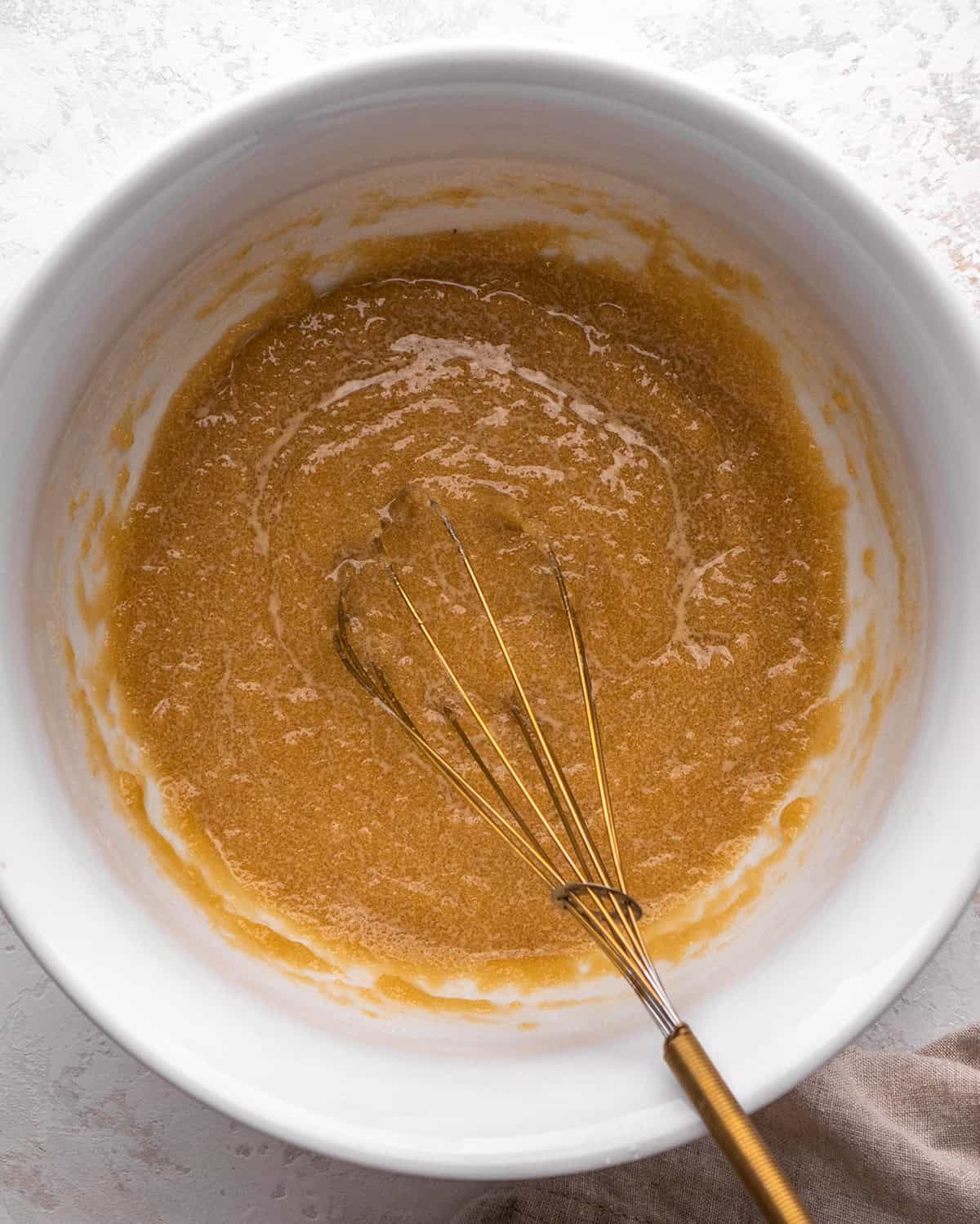 how to make blondies - melted butter, white sugar and brown sugar whisked together in a white bowl with a gold whisk