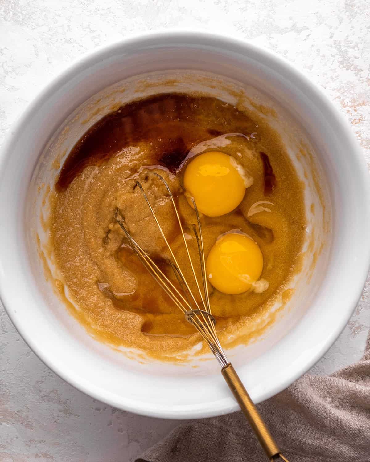 how to make blondies - eggs and vanilla added to the melted butter & sugar mixture in a white bowl before mixing, there is a gold whisk int eh bowl.
