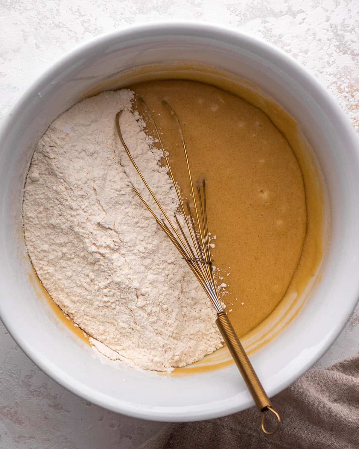 how to make blondies - dry ingredient mixture added to the wet ingredient mixture in a white bowl with a gold whisk before mixing