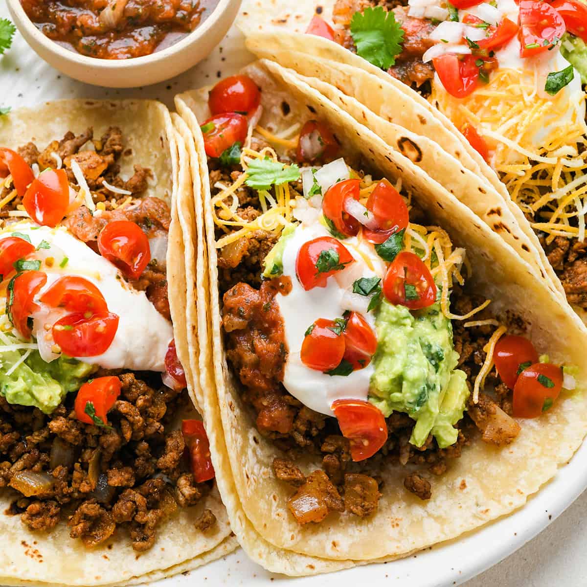 3 Ground Beef Tacos on a plate with sour cream, cheese, guacamole and tomatoes
