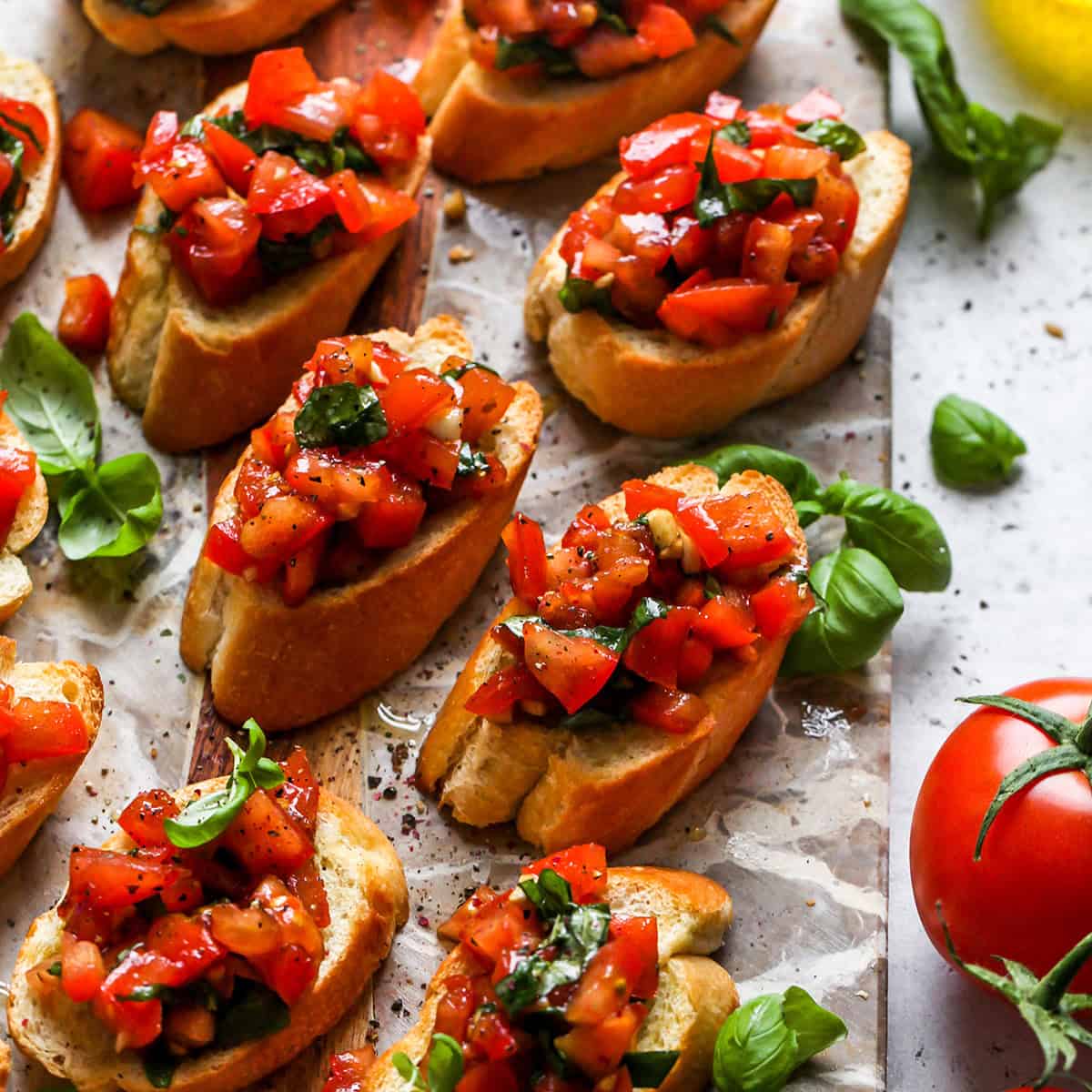 7 pieces of toasted bread topped with this easy tomato bruschetta recipe with diced tomatoes and fresh basil, served on a marble and wood serving board surrounded by fresh basil leaves