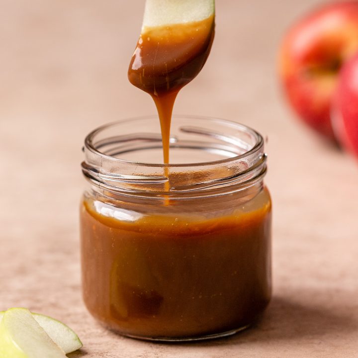 a glass jar of Caramel Apple Dip and an apple being held over the jar after being dipped inside