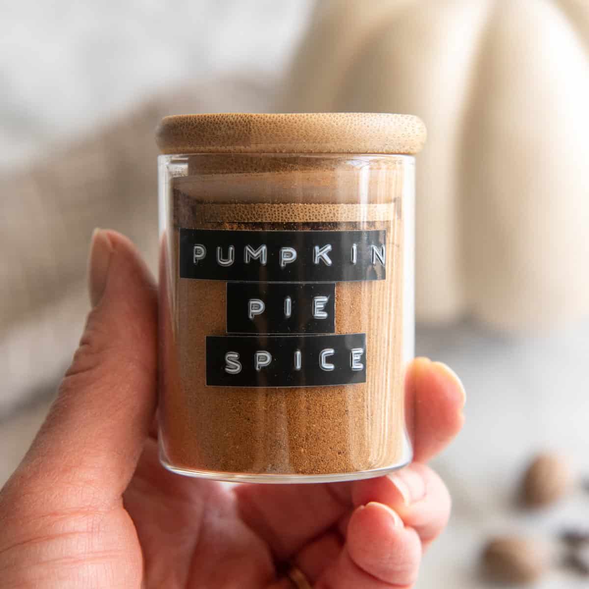 a hand holding a glass jar of Homemade Pumpkin Pie Spice with a wooden lid and a black label 